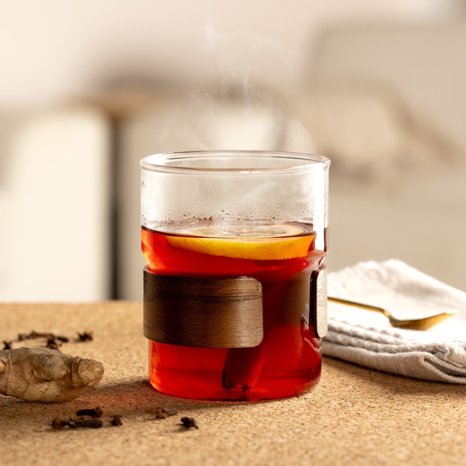 Ein dekoratives Teeglas mit schwarzem Tee, Zitronenscheibe und Zimtstange, serviert auf einem Korkuntergrund. Im Vordergrund liegen Gewürznelken und ein Stück frischer Ingwer.