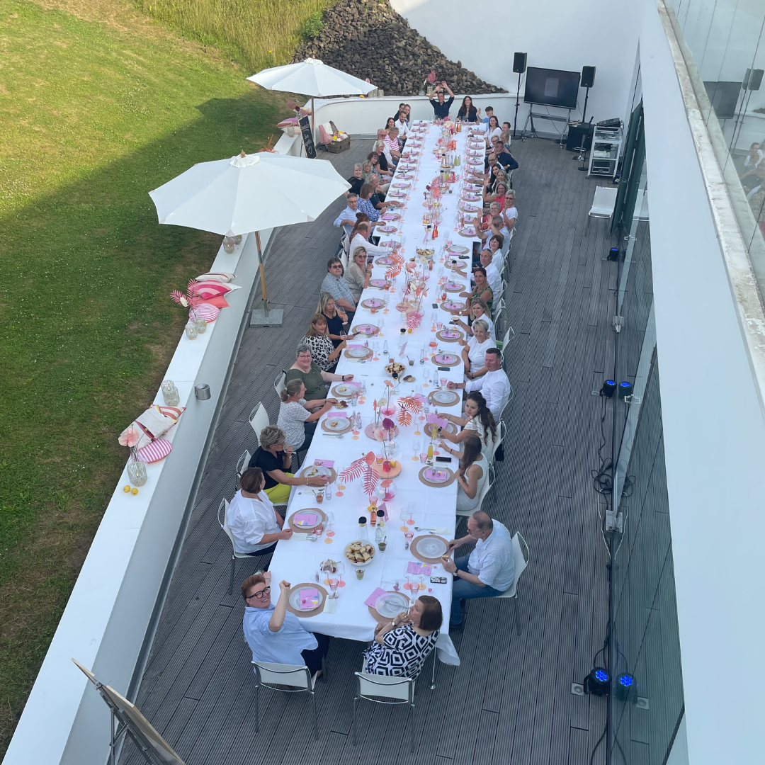 Langes festlich gedecktes Dinner unter freiem Himmel mit vielen fröhlichen Gästen an einer weiß gedeckten Tafel.