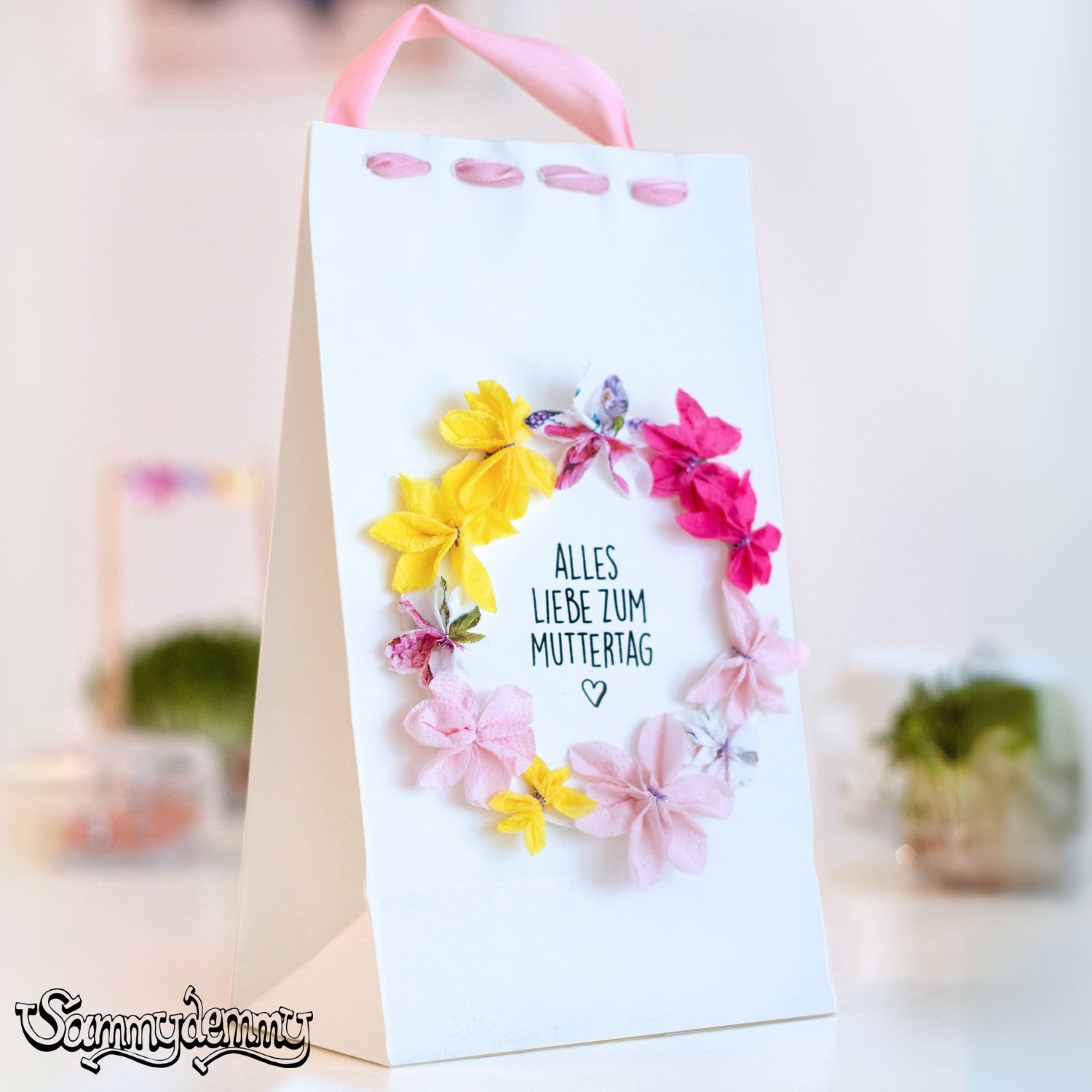 Eine liebevoll gestaltete weiße Geschenktüte mit einem Herz aus bunten Stoffblumen in Rosa, Gelb und Pink. In der Mitte steht der handgeschriebene Schriftzug „Alles Liebe zum Muttertag“. Die rosa Schleife am oberen Rand rundet das Design charmant ab.