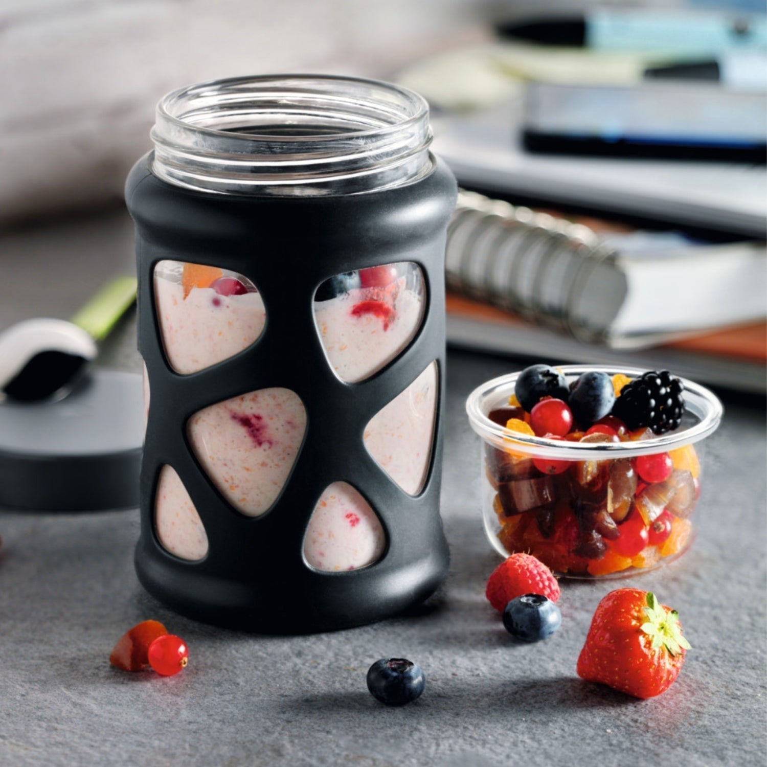 Joghurt mit Beeren in einem Glas mit schwarzer Silikonhülle – daneben frische Früchte in einem kleinen Glas.