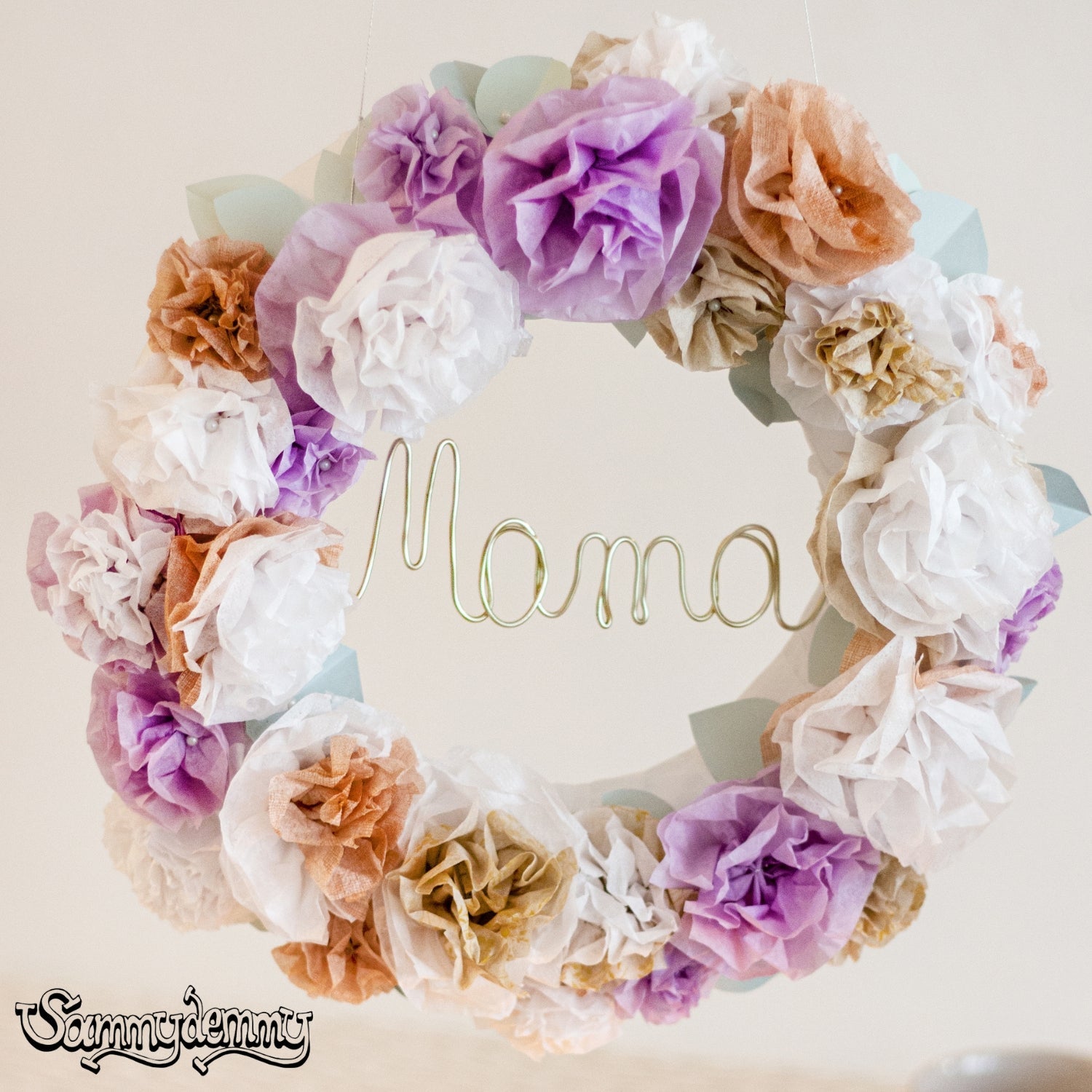 Ein liebevoll gestalteter Blumenkranz aus Seidenpapierblüten in Weiß, Lila, Beige und Apricot hängt an einem Faden. In der Mitte ist das Wort „Mama“ aus goldfarbenem Draht geformt – eine kreative DIY-Idee zum Muttertag oder als persönliches Geschenk