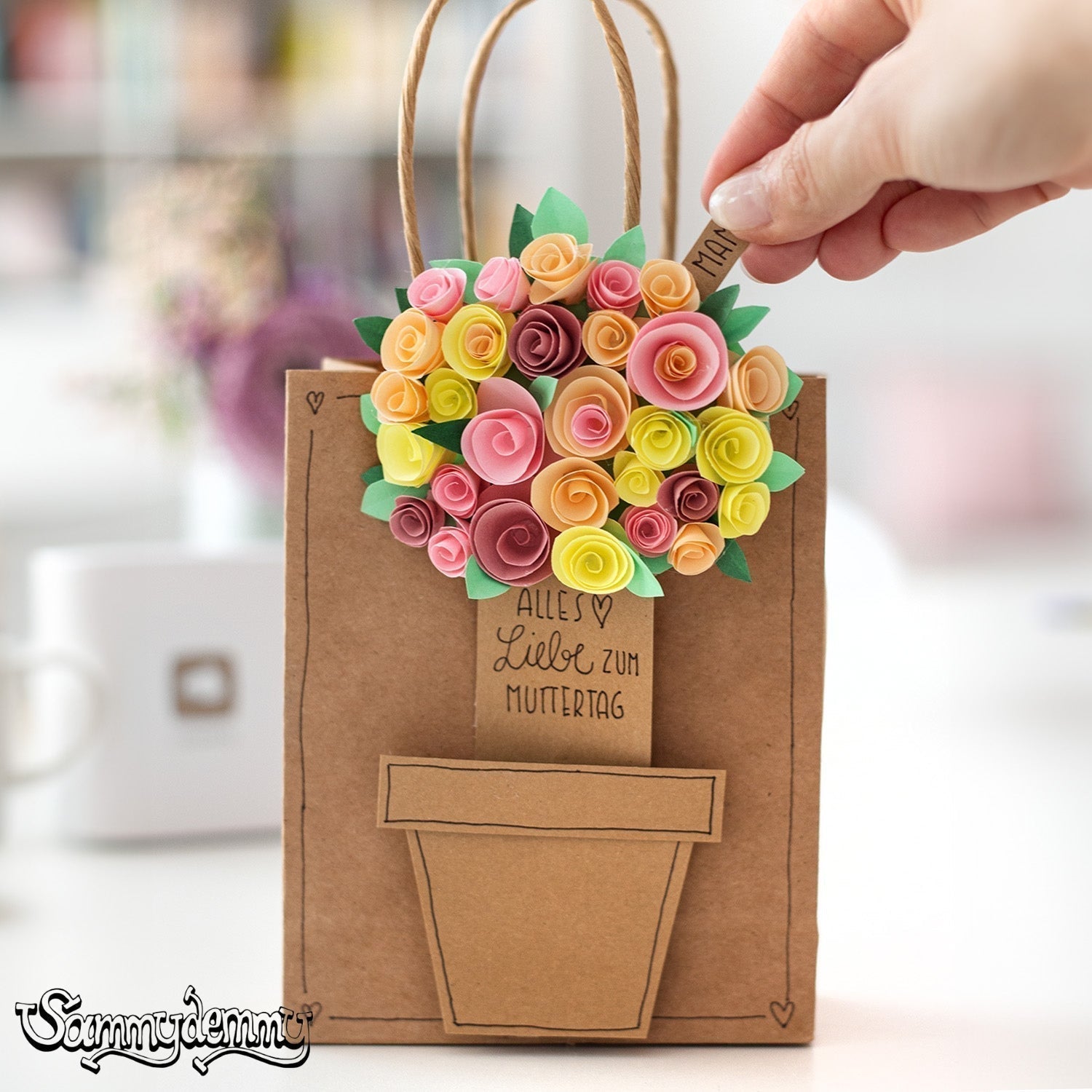 Eine braune Geschenktüte mit aufgeklebtem Blumentopf und einem kunstvollen Blumenstrauß aus buntem Papier – in der Mitte steht „Alles Liebe zum Muttertag“. Eine Hand zieht einen kleinen Anhänger mit der Aufschrift „Mama“ heraus