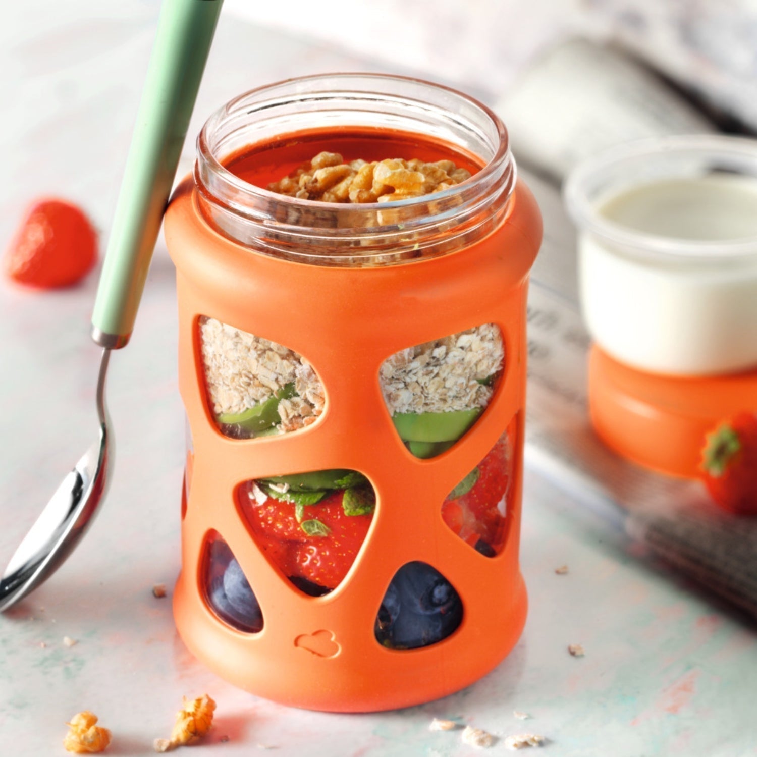 Schichtglas mit Haferflocken, Früchten und Nüssen in orangener Silikonhülle – daneben ein Becher Joghurt.