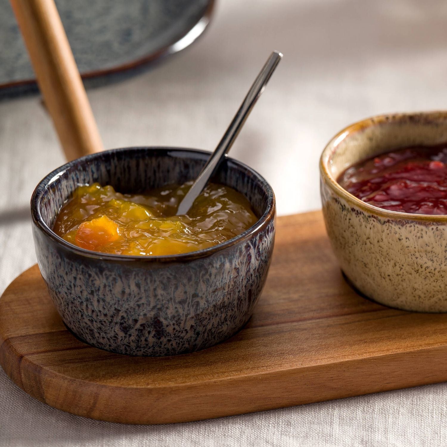Zwei kleine Keramikschalen von LEONARDO auf einem Holzbrett – eine gefüllt mit gelber Marmelade samt Löffel, die andere mit rotem Fruchtaufstrich.