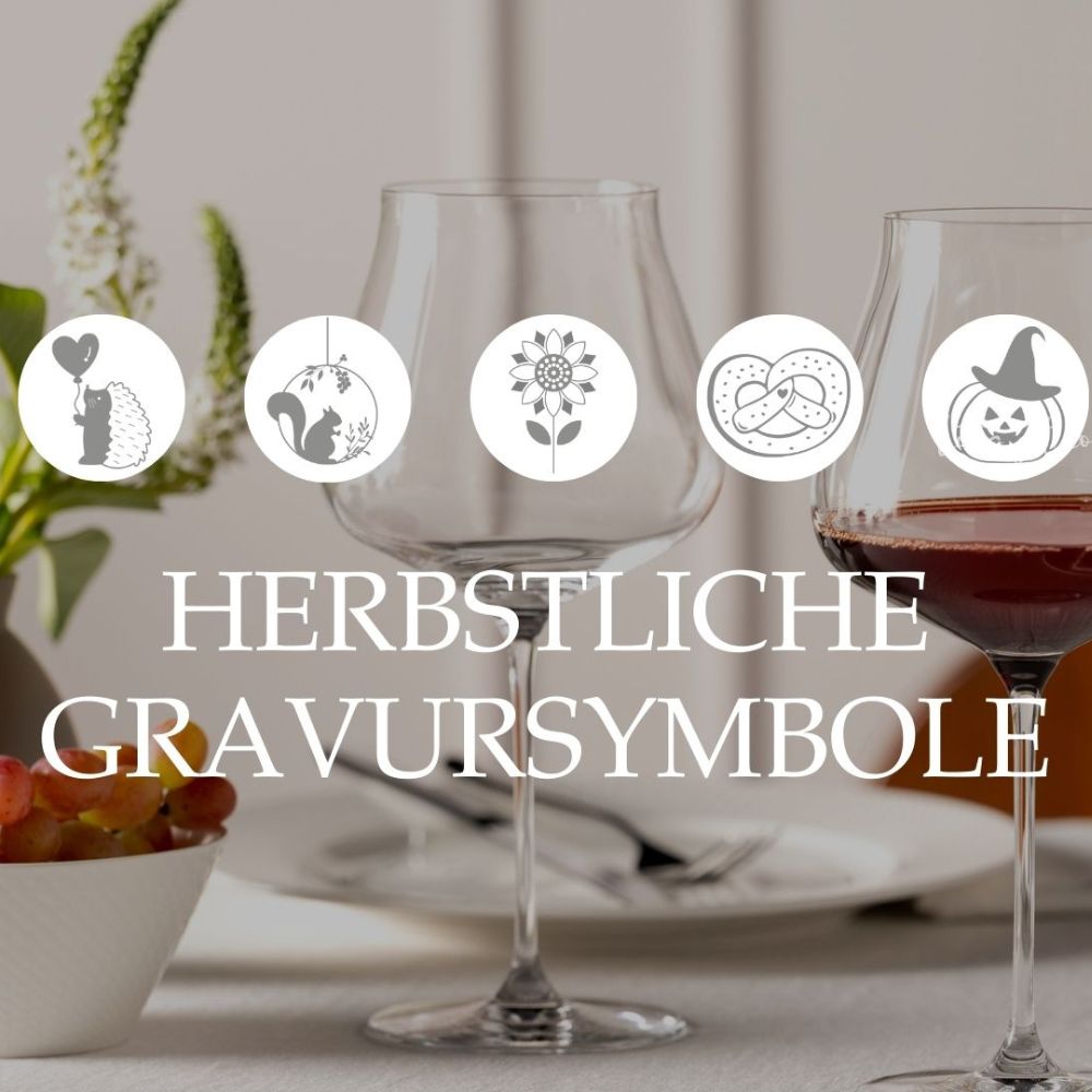 Zwei Weingläser mit herbstlichen Gravursymbolen, im Hintergrund herbstlich gedeckter Tisch mit Blumen und Trauben.
