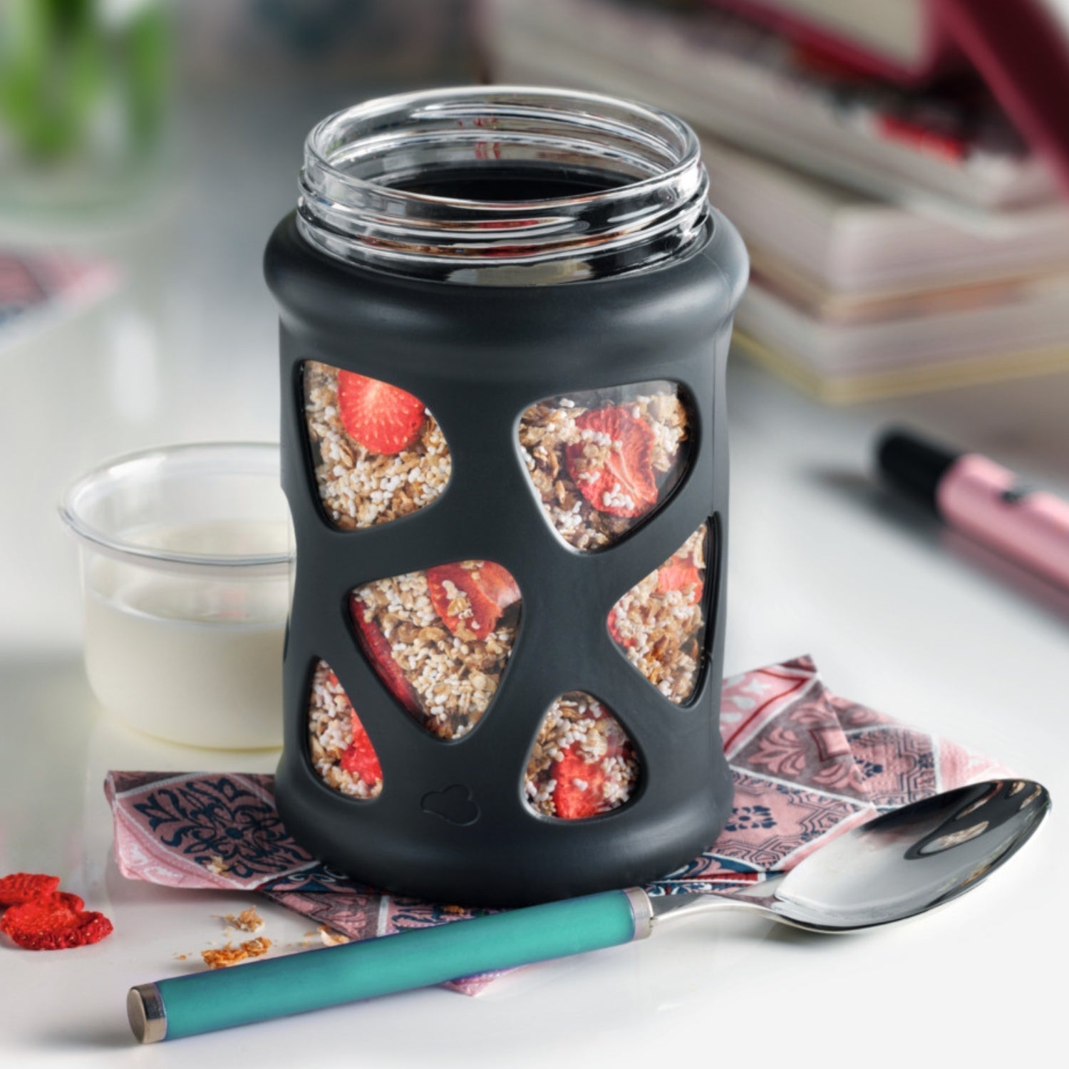 Müsli mit getrockneten Erdbeeren im schwarzen Glas – daneben ein kleiner Becher mit Milch oder Joghurt