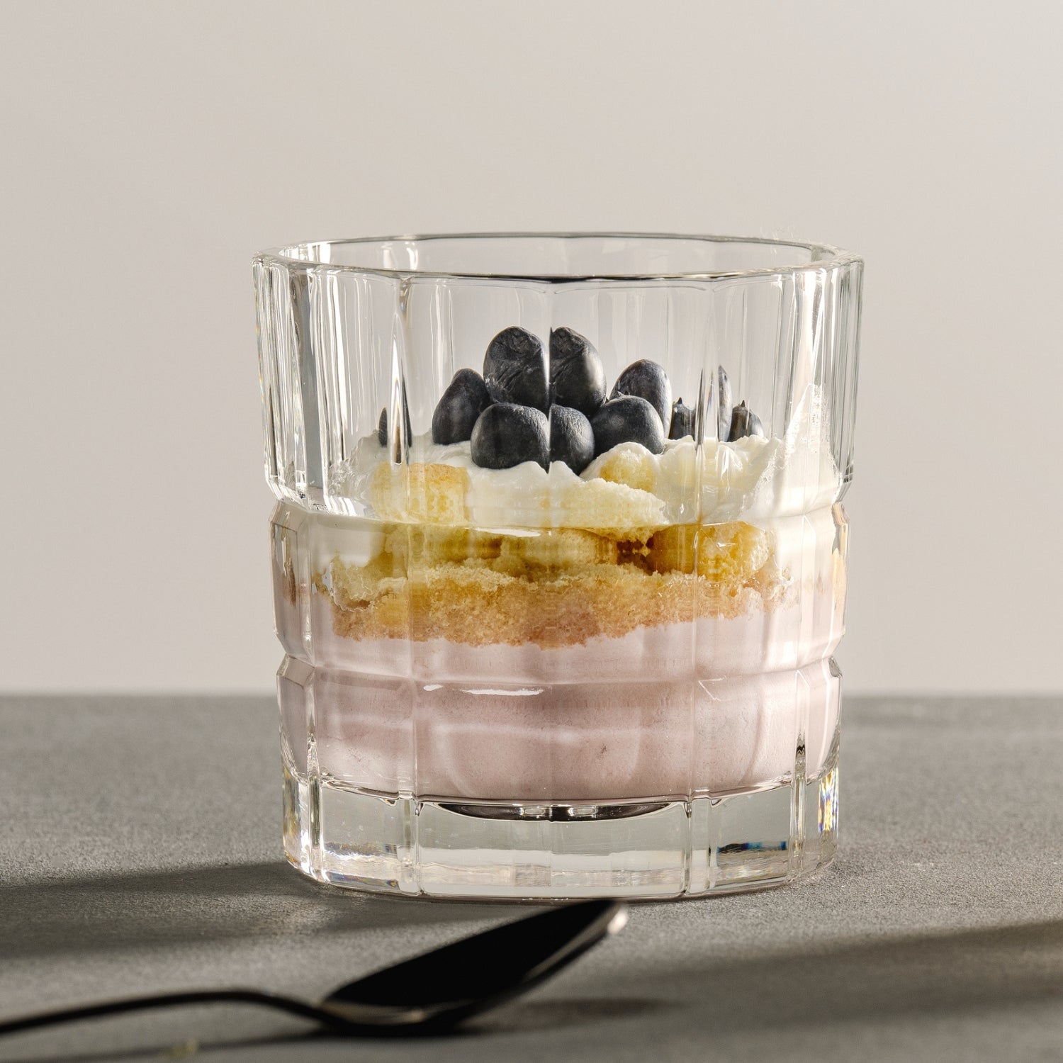 Dessert im Kristallglas mit Schichten aus Beerencreme, Biskuit, Sahne und frischen Heidelbeeren auf grauer Oberfläche.