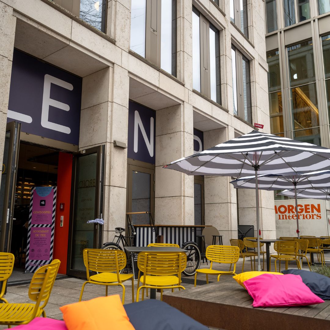 LEONARDO Cafe in Frankfurt am Main. Außenansicht eines modernen Gebäudes mit großen Fenstern, gelben Stühlen und gestreiften Sonnenschirmen vor dem Eingang eines Cafés.