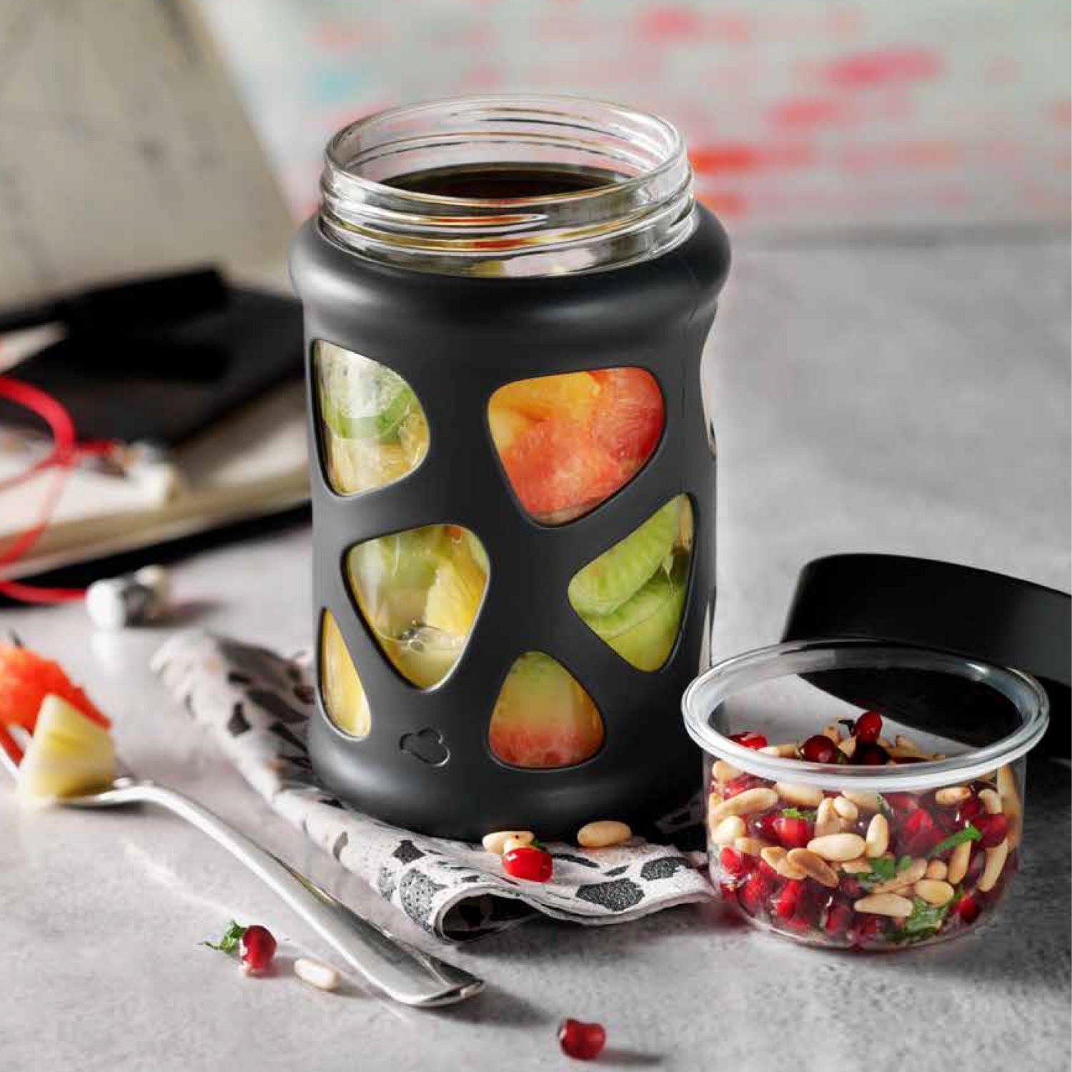 Obstsalat in Glas mit schwarzer Silikonhülle – daneben ein Glas mit Pinienkernen und Granatapfelkernen.