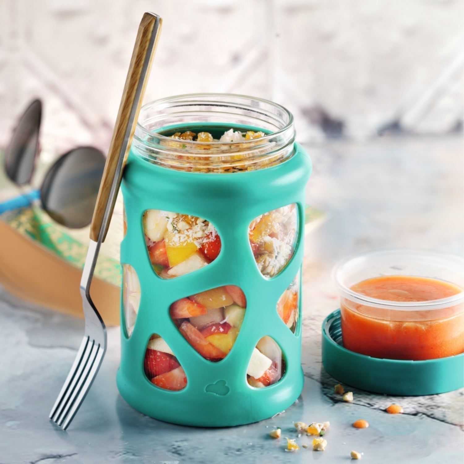 Bunter Obstsalat im Glas mit türkisfarbener Silikonhülle und Dipdöschen.