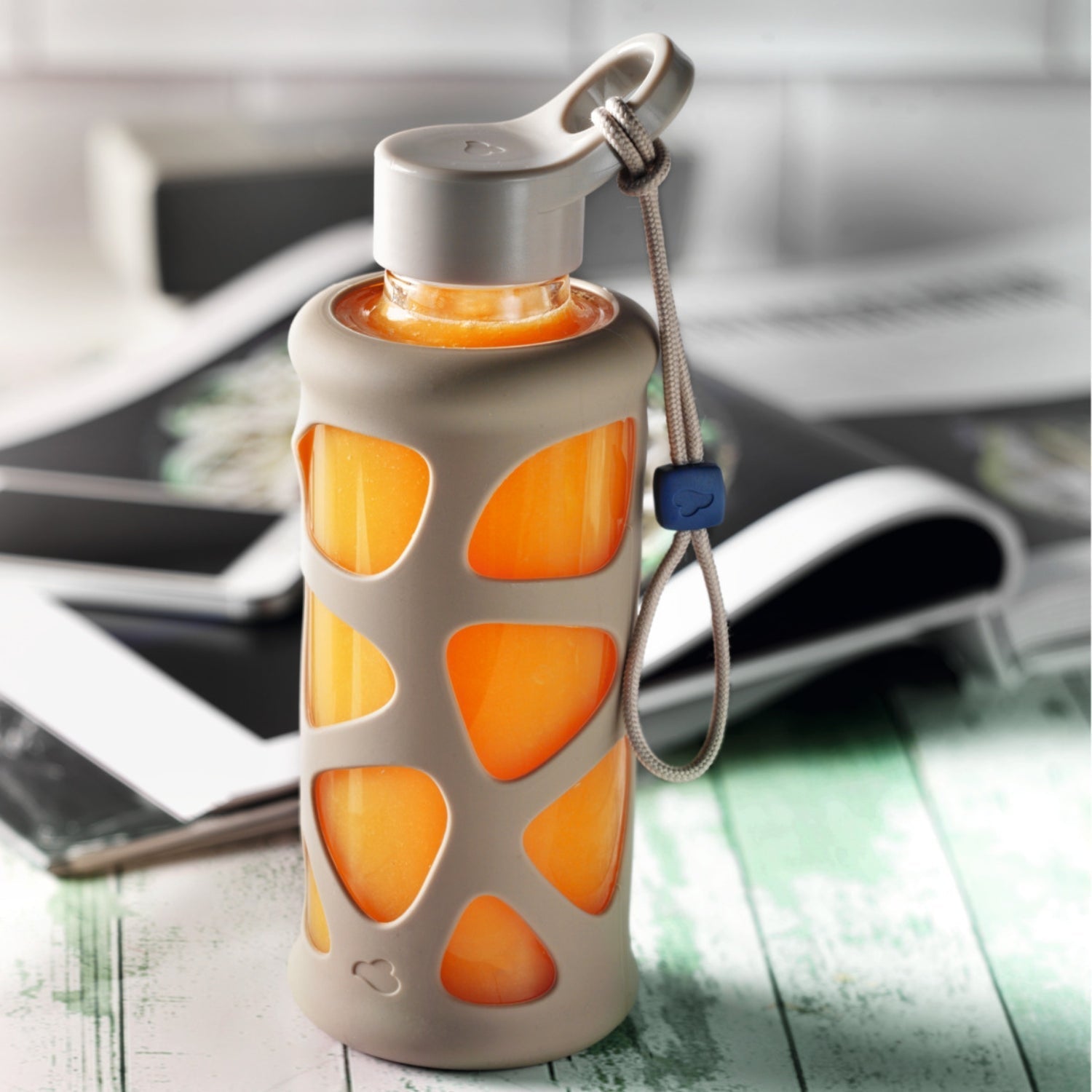 Kompakte Glasflasche mit orangem Getränk und beiger Hülle, auf Magazin liegend