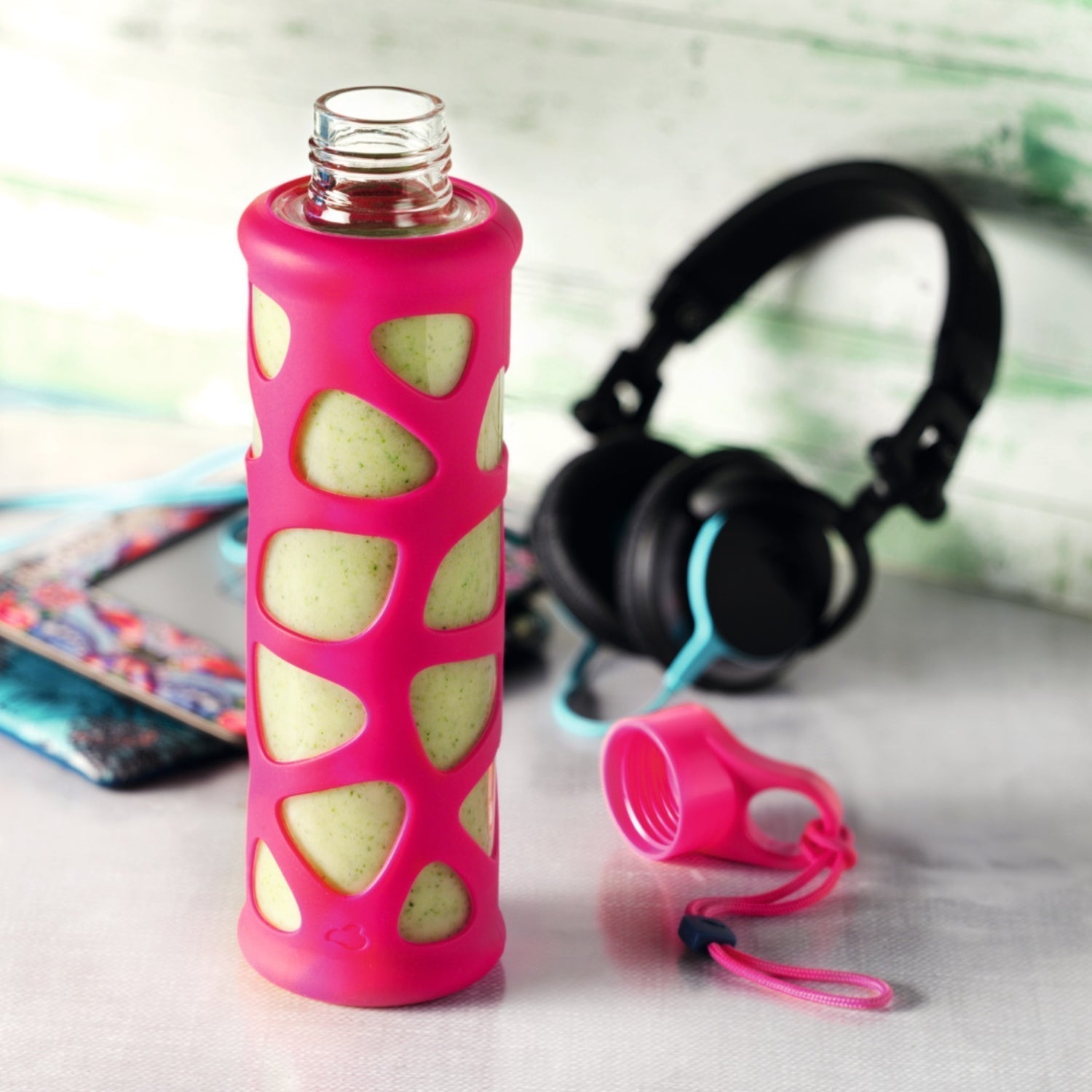 Glasflasche mit grünem Smoothie und pinker Silikonhülle vor Kopfhörern