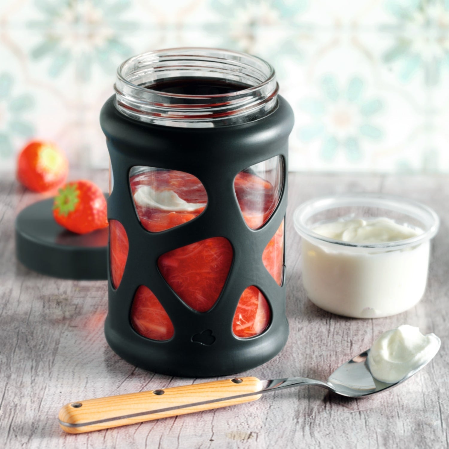 Fruchtiges Schichtdessert mit Erdbeeren im Glas mit schwarzer Hülle – daneben ein Löffel mit Quark.