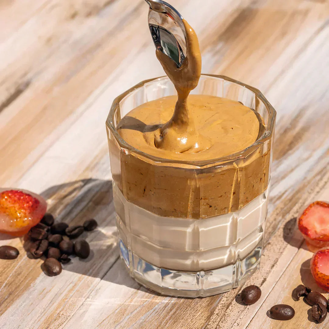 Cremiger Dalgona-Kaffee auf Milch im Facettenglas, umgeben von Kaffeebohnen und Erdbeeren.