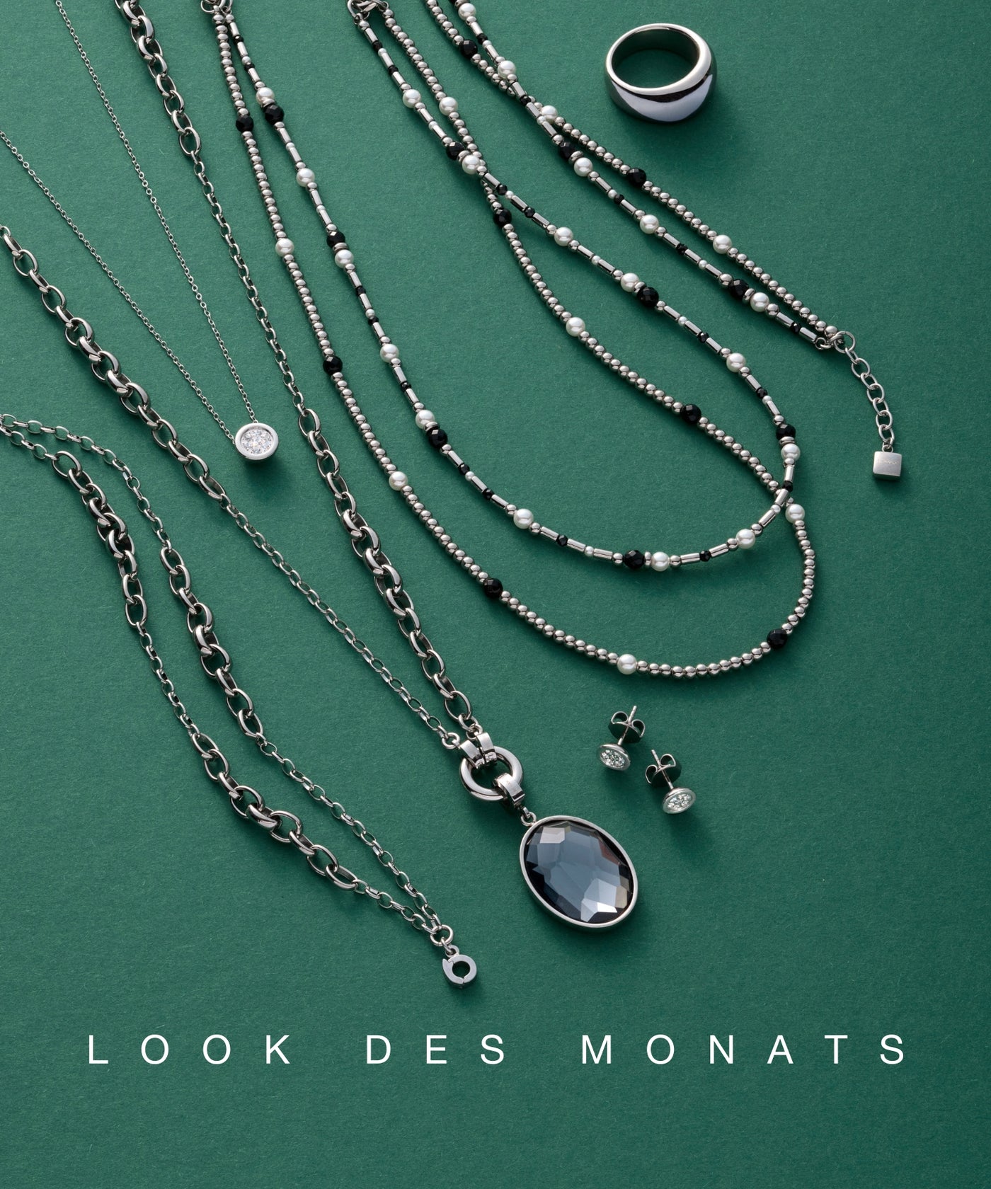 Ein Arrangement aus silberfarbenem und schwarz-weißen Schmuckstücken – darunter Halsketten, Ohrstecker und ein Ring – liegt auf grünem Hintergrund mit dem Titel „LOOK DES MONATS“.