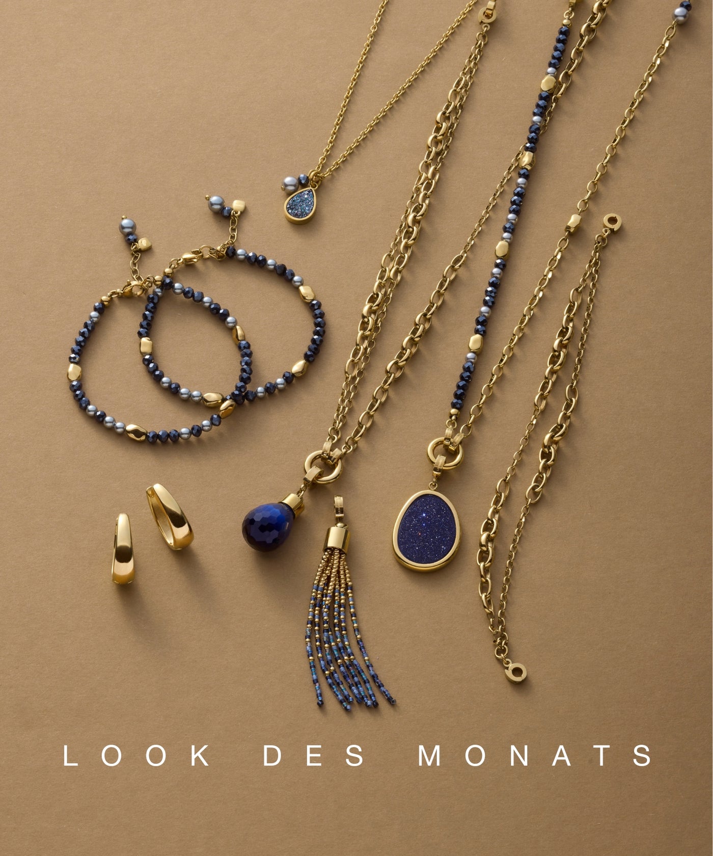 Goldfarbene Schmuckstücke mit blauen Akzenten – darunter Ketten, Armbänder, Ohrstecker und Anhänger – sind auf einem warmen, braunen Untergrund stilvoll arrangiert; unten steht „LOOK DES MONATS“.