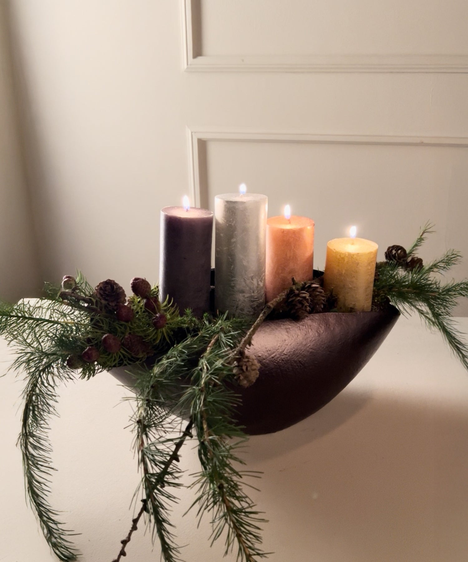 Adventsgesteck mit vier brennenden Stumpenkerzen in Lila, Silber, Orange und Gelb, dekoriert mit Tannenzweigen und Zapfen in einer braunen Schale.