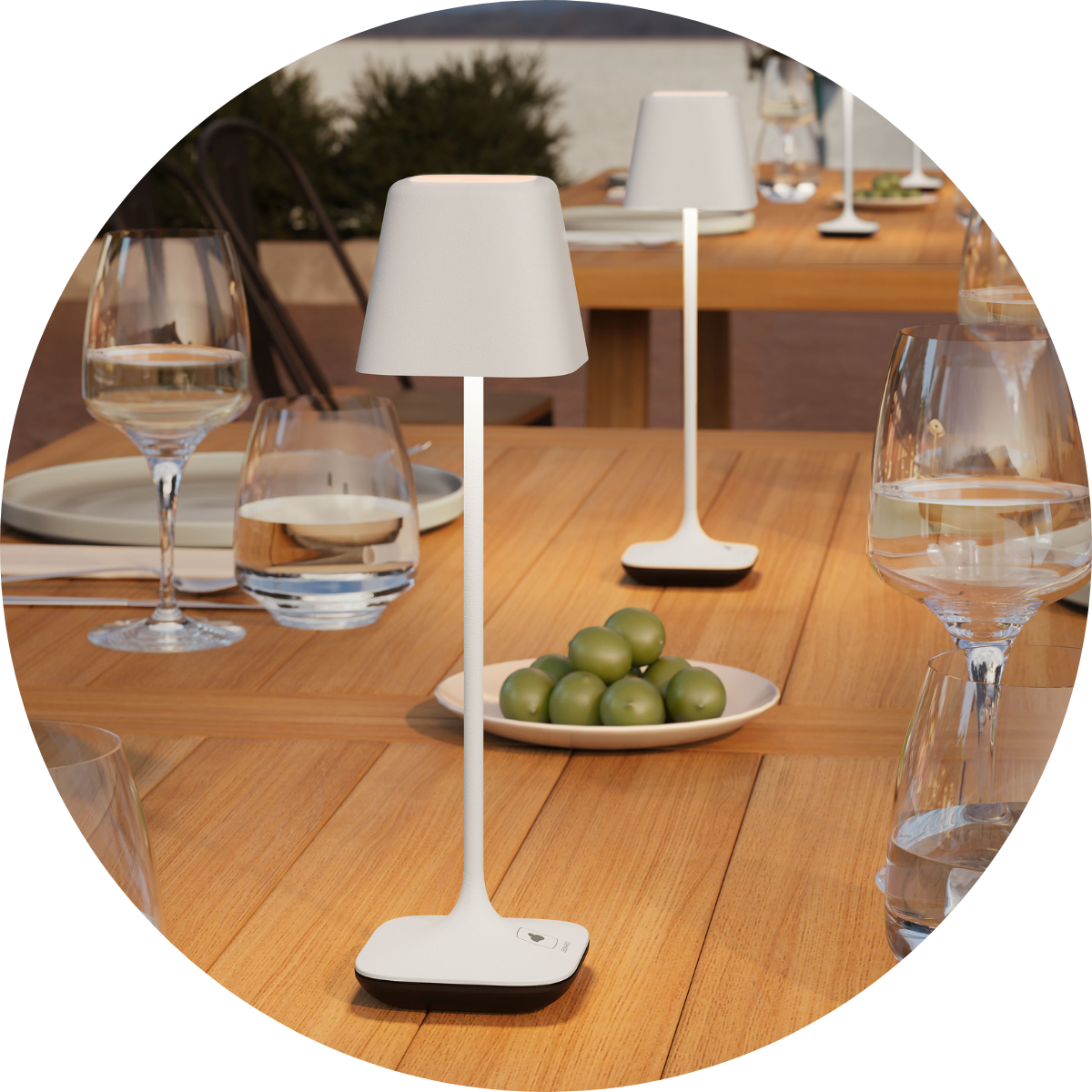 Elegante weiße Akku-Tischleuchten stehen auf einem gedeckten Holztisch im Außenbereich mit Gläsern, Tellern und einer Schale Oliven.