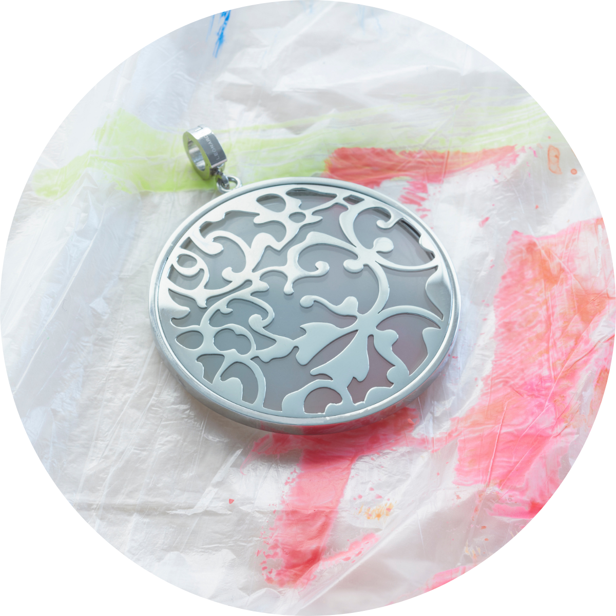 Silberfarbener Anhänger mit verschnörkeltem Blumenmuster liegt auf einer transparenten Unterlage mit bunten Farbklecksen.