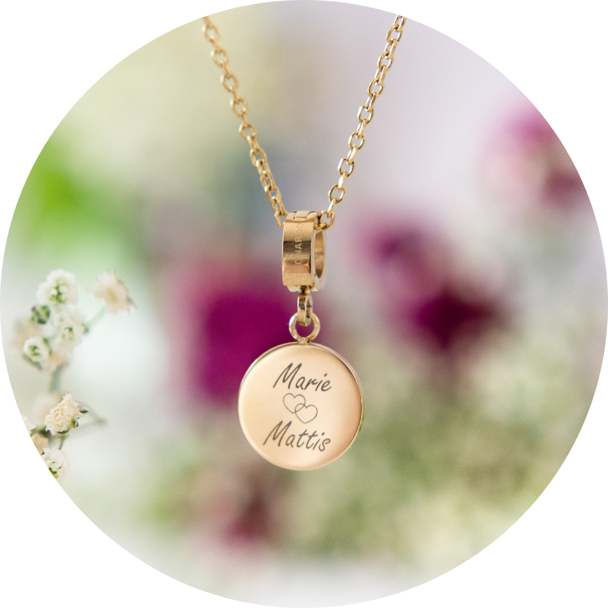Feine goldene Kette mit rundem, goldenem Anhänger, graviert mit den Namen „Marie“ und „Mattis“ und einem kleinen Herzsymbol, vor blumigem Hintergrund.