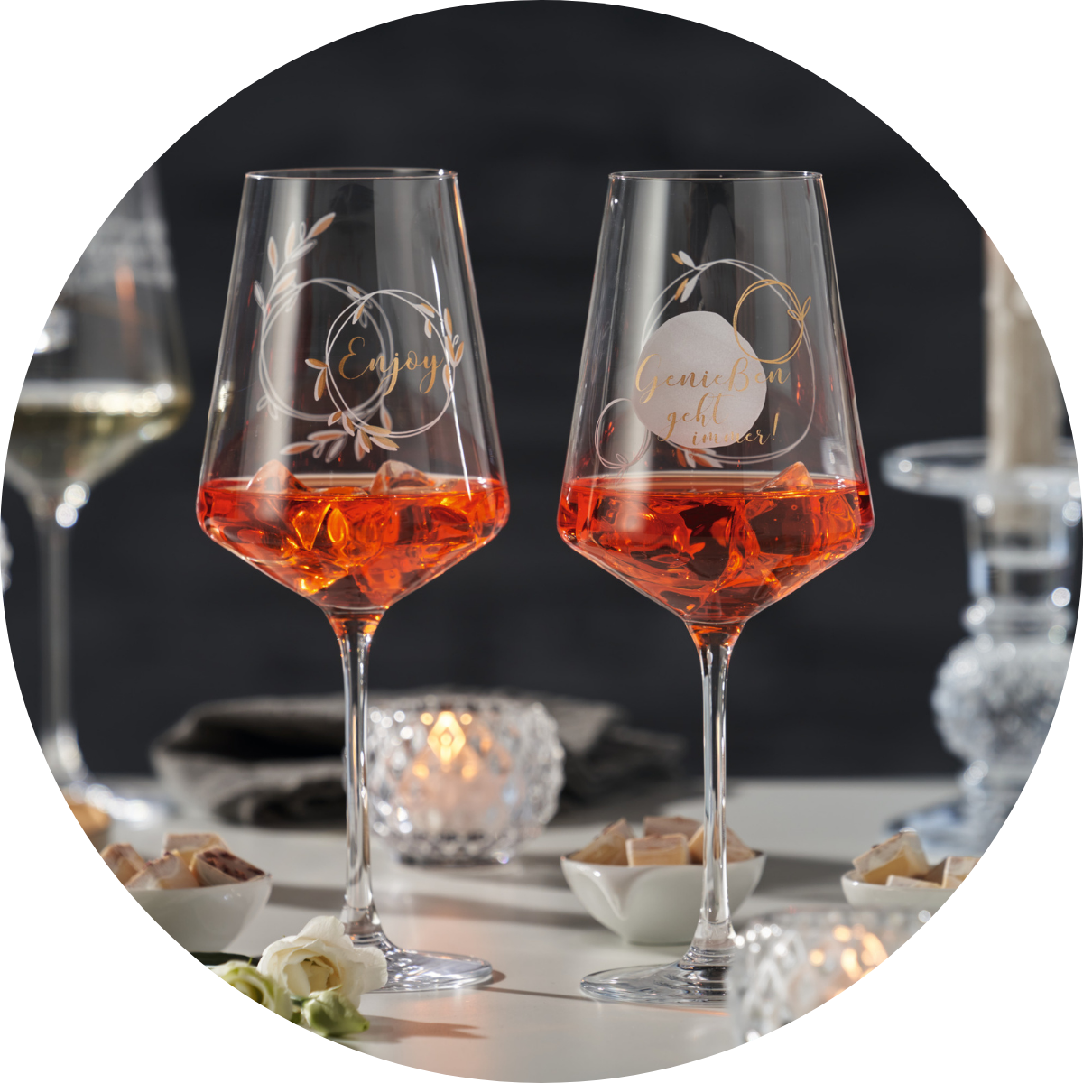 Elegante Aperitifgläser mit Gravur „Enjoy“ und „Genießen geht immer“ auf einem gedeckten Tisch mit Kerzenlicht und Snacks.