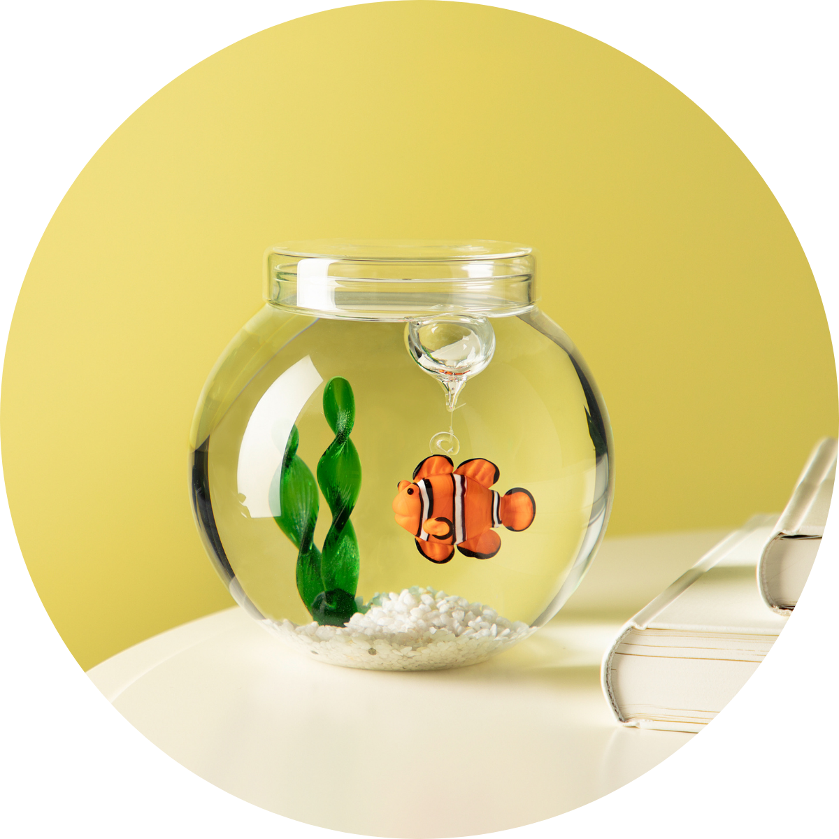 Kleine Glaskugel mit handgefertigtem Clownfisch und grüner Glas-Pflanze auf weißen Steinen, platziert auf einem Tisch vor gelbem Hintergrund.