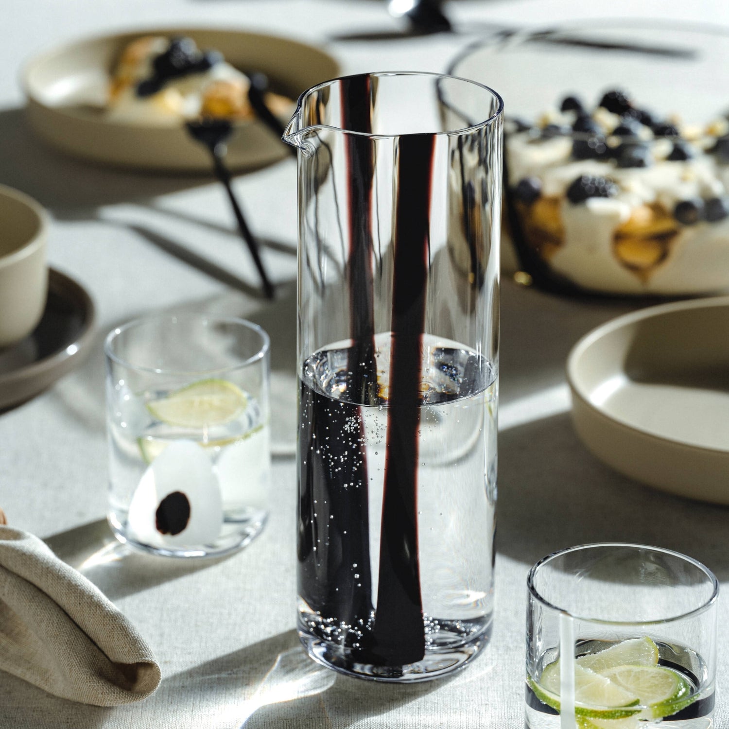 Eine elegante Glaskaraffe mit auffälligem schwarz-rotem Dekor steht im Mittelpunkt einer stilvoll gedeckten Tafel und ist mit sprudelndem Wasser gefüllt – umgeben von Gläsern mit Limettenscheiben und einem sommerlichen Dessert.