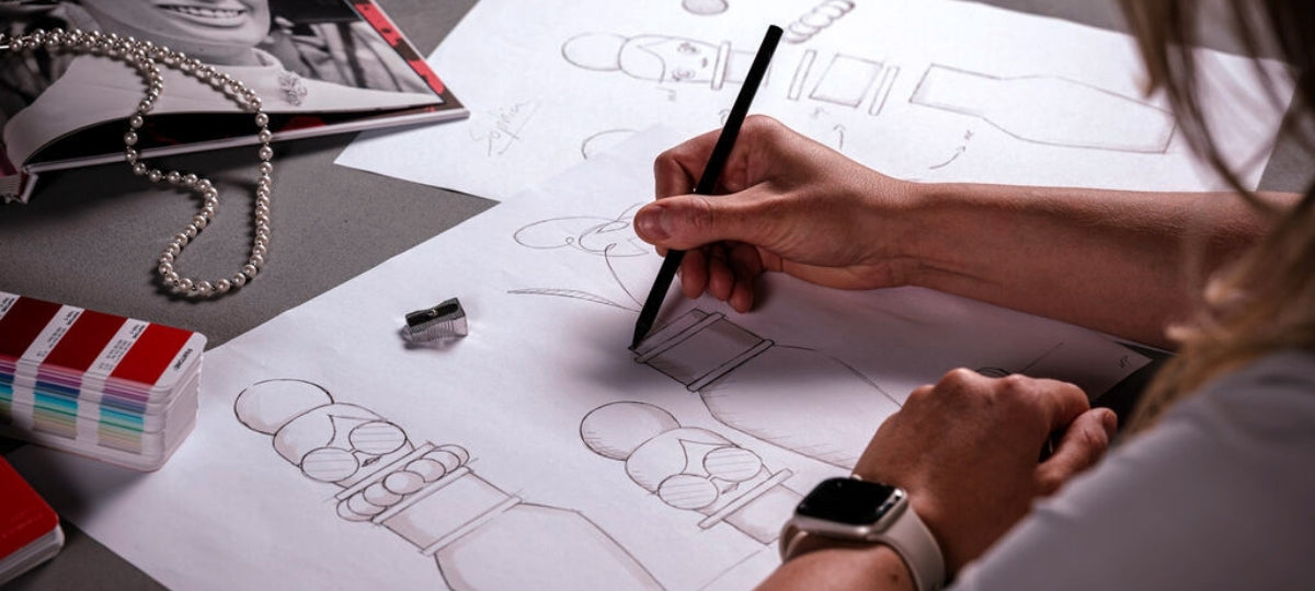 Eine Person zeichnet mit einem schwarzen Stift Figurenentwürfe auf Papier. Neben ihr liegen Farbkarten, ein Spitzer und eine Perlenkette.