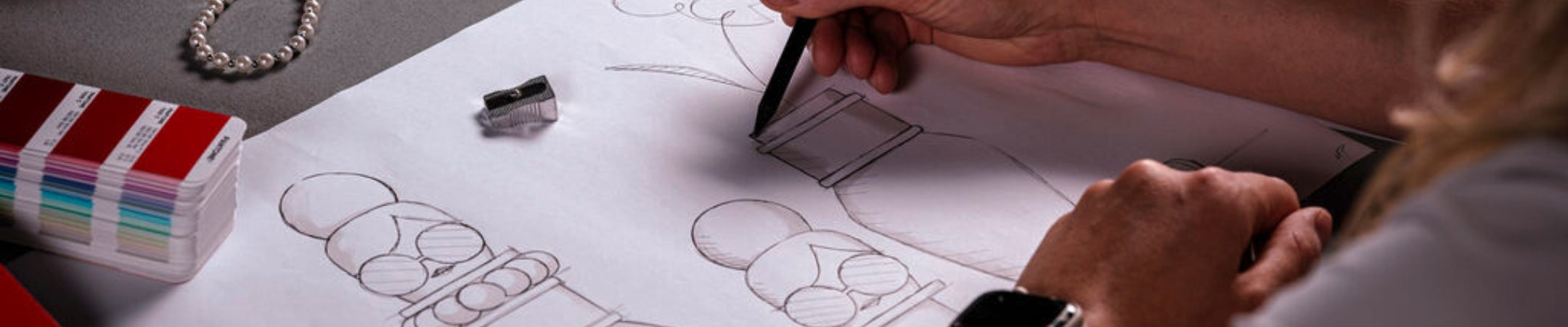 Detailaufnahme einer Hand, die eine stilisierte Figur auf Papier skizziert. Links liegen Farbkarten und eine Perlenkette.