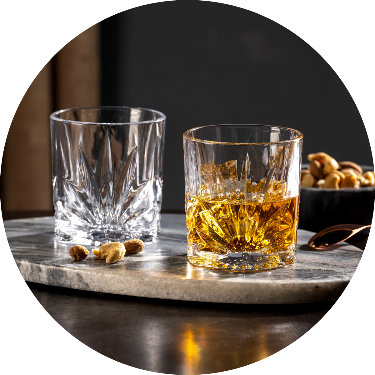 Zwei elegante Bargläser, eines leer und eines mit Whisky und Eis, auf einer Marmorplatte mit Nüssen daneben.