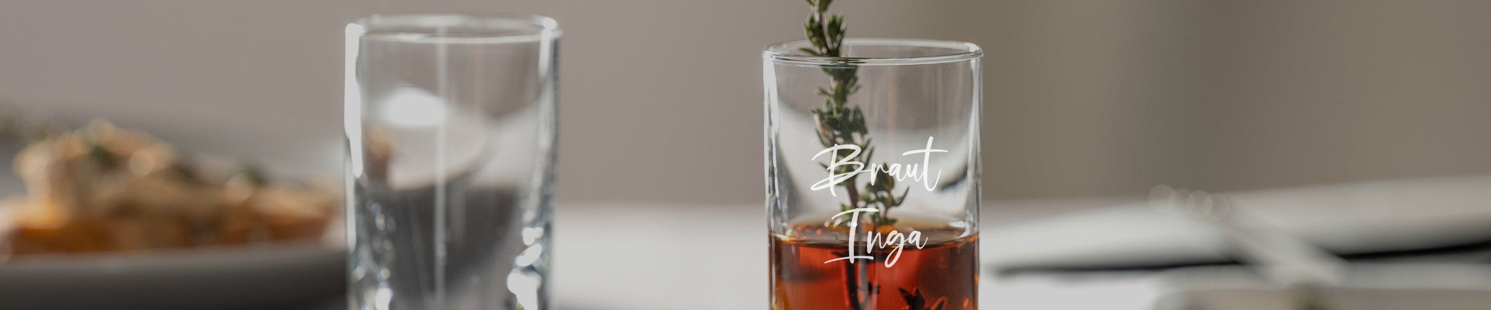 Zwei LEONARDO Shotgläser, eines davon graviert mit „Braut Inga“, dekoriert mit Thymianzweig, auf gedecktem Tisch mit unscharfem Hintergrund.
