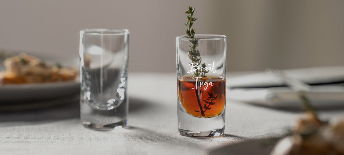 Zwei LEONARDO Shotgläser, eines davon graviert mit „Braut Inga“, dekoriert mit Thymianzweig, auf gedecktem Tisch mit unscharfem Hintergrund.