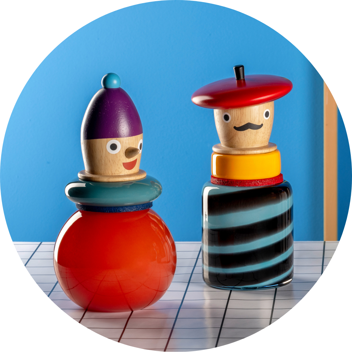 Zwei bunte Vasen mit Holzfiguren-Köpfe als Aufsätze stehen auf einer weiß-blauen Tischplatte vor blauem Hintergrund.