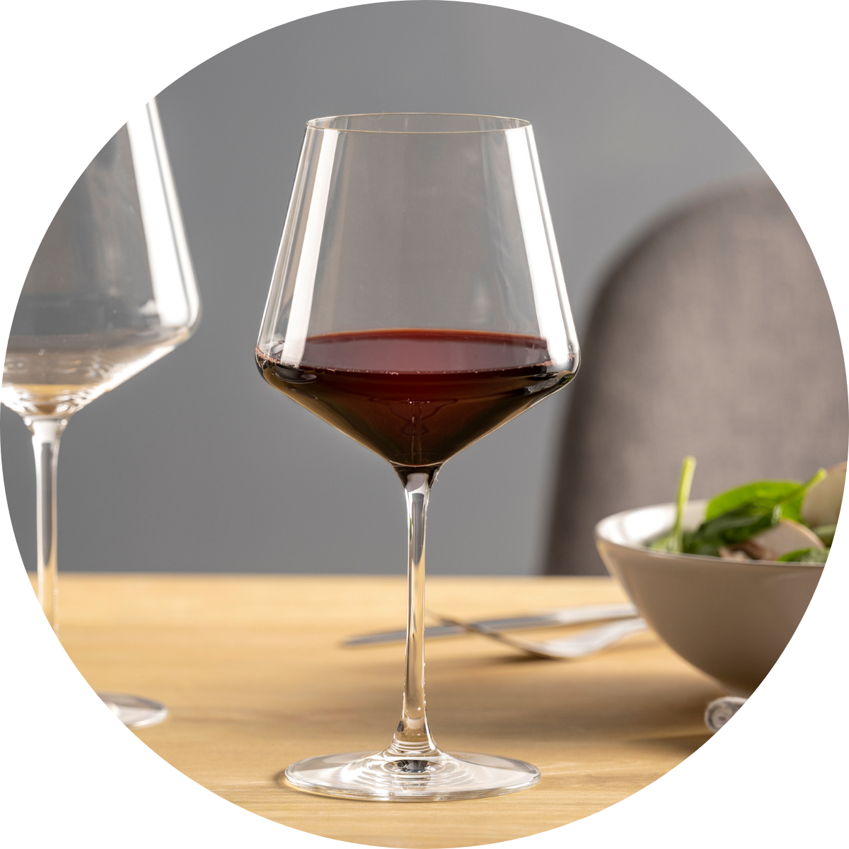 Burgunderglas mit Rotwein neben einem Salatteller auf hellem Holztisch.