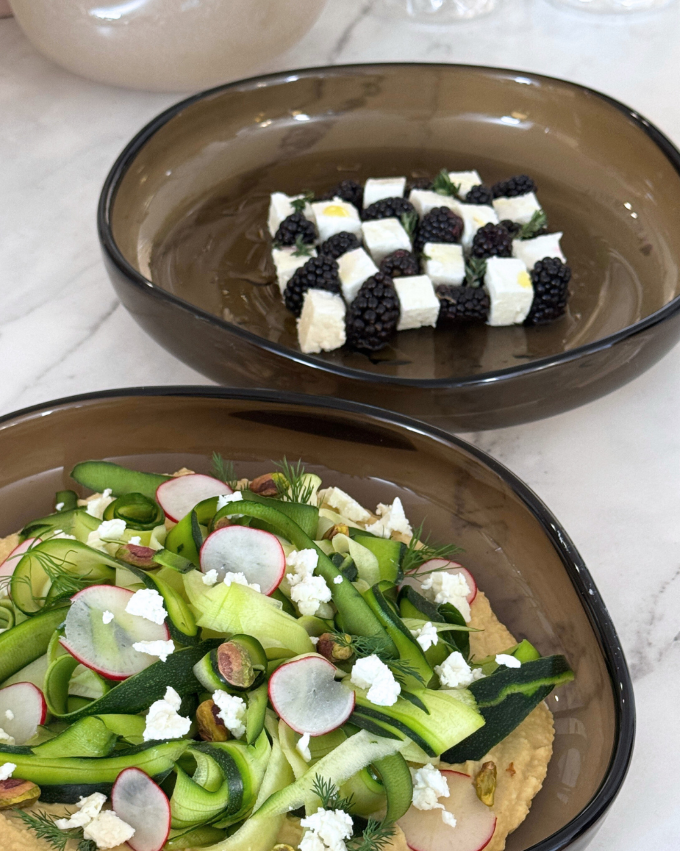 Zwei braune Glasschalen mit Salaten, unter anderem mit Zucchinistreifen, Radieschen, Feta und Brombeeren.