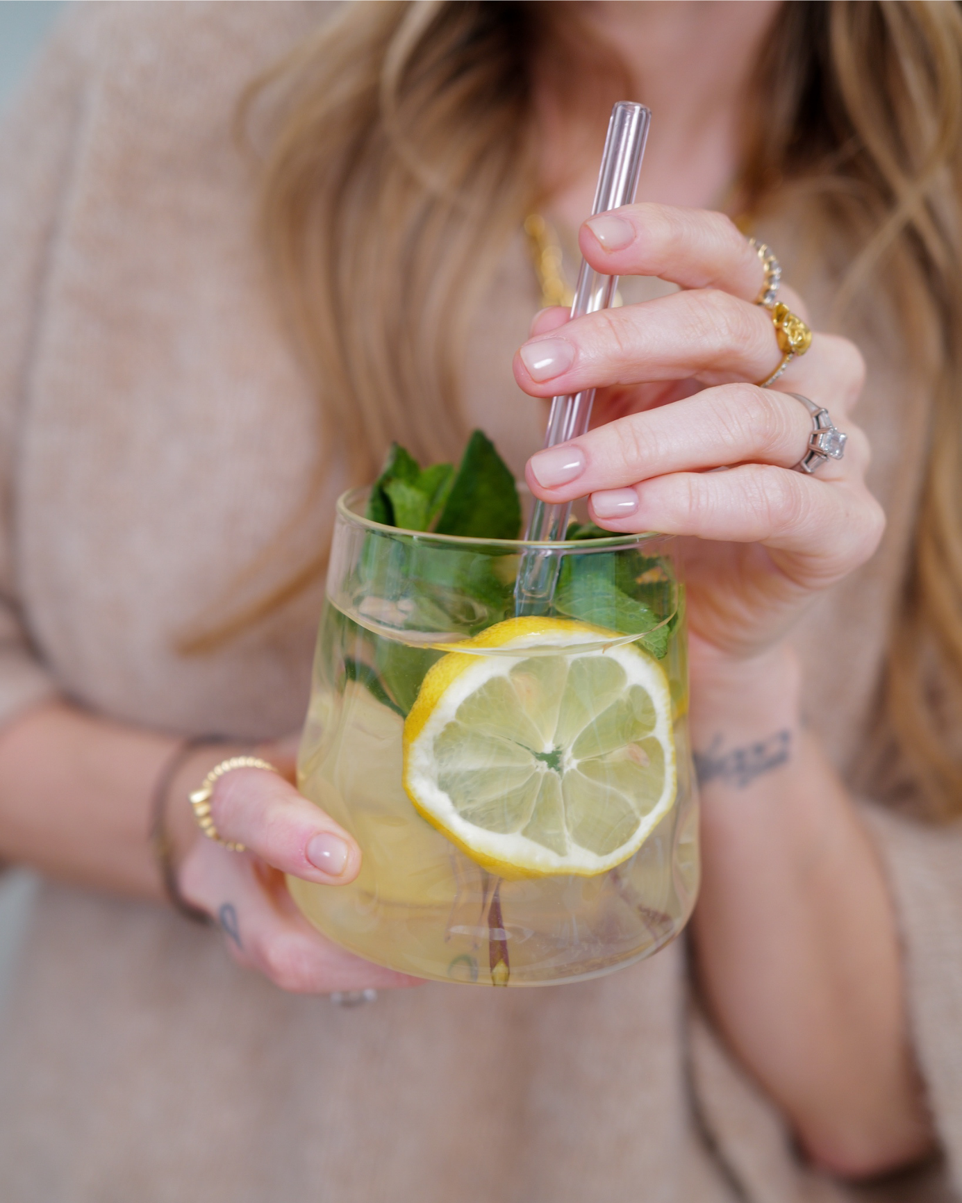 Nahaufnahme eines Glasbechers mit Zitronenscheibe und Minze, eine Person hält einen Glastrinkhalm.