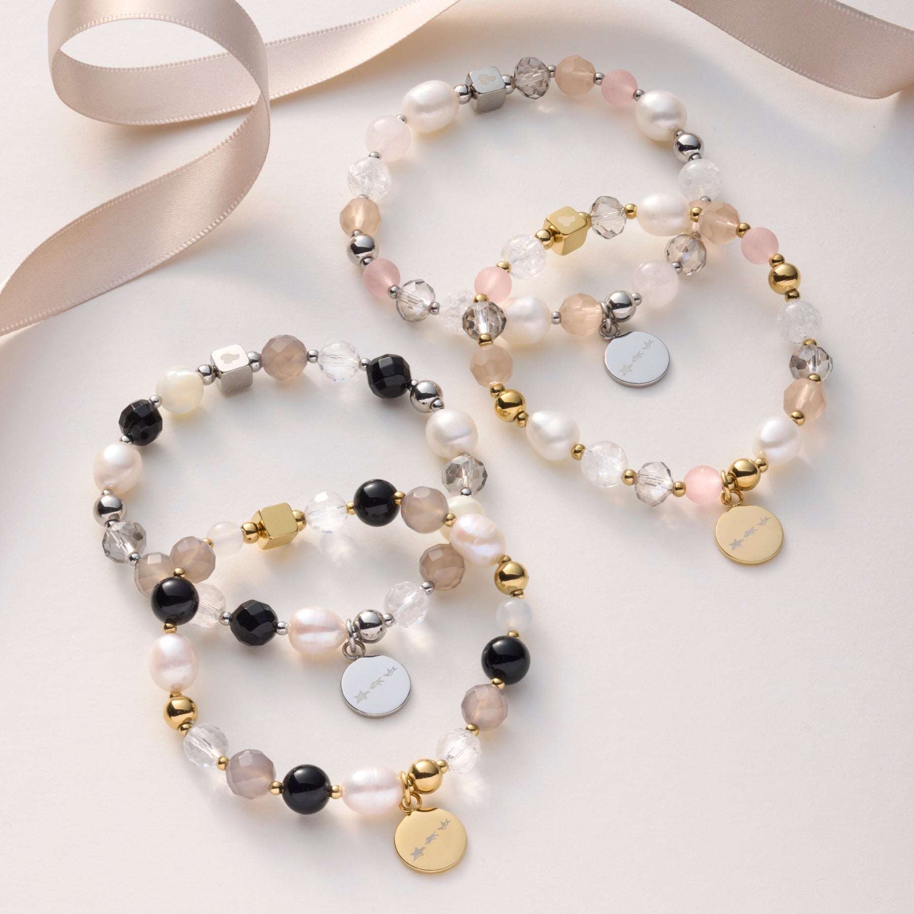 Vier Perlenarmbänder in zarten Nude-, Weiß- und Schwarztönen mit funkelnden Steinen und goldenen sowie silbernen Plaketten liegen dekorativ neben einem Satinband.