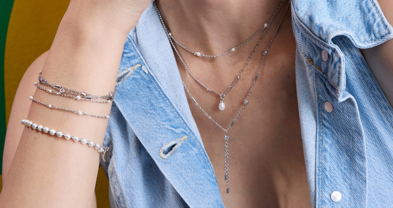 Frau im Jeanshemd mit mehreren filigranen Ketten in Silberoptik und Perlendetails sowie passenden Armbändern am Handgelenk; Fokus auf Dekolleté und Schmuck vor farbigem Hintergrund.