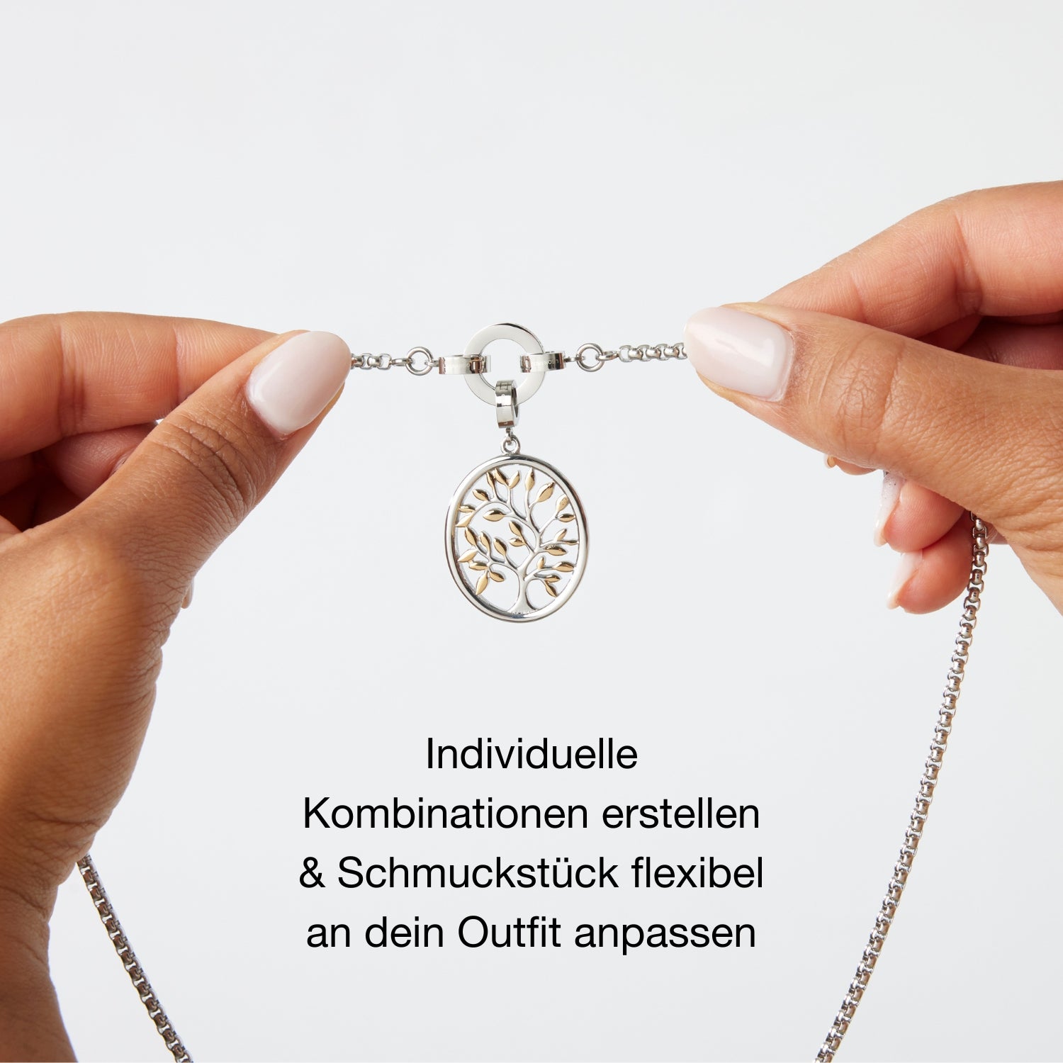 Zwei Hände halten eine Halskette mit Clip-Verschluss, an dem ein runder Lebensbaum-Anhänger befestigt ist. Darunter steht: „Individuelle Kombinationen erstellen & Schmuckstück flexibel an dein Outfit anpassen“.