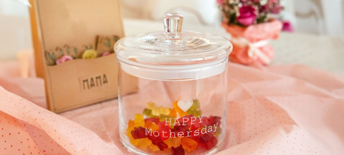 LEONARDO Glasdose TOP mit Gravur „HAPPY Mothersday“, gefüllt mit Gummibärchen, dekorativ inszeniert mit Muttertagsgeschenk im Hintergrund.