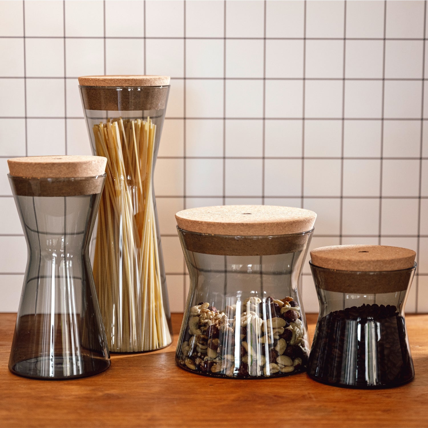 Vier formschöne Vorratsgläser aus Glas mit natürlichem Korkdeckel stehen auf einer Holzfläche vor gefliestem Hintergrund und sind stilvoll mit Pasta, Nüssen und Kaffeebohnen befüllt.
