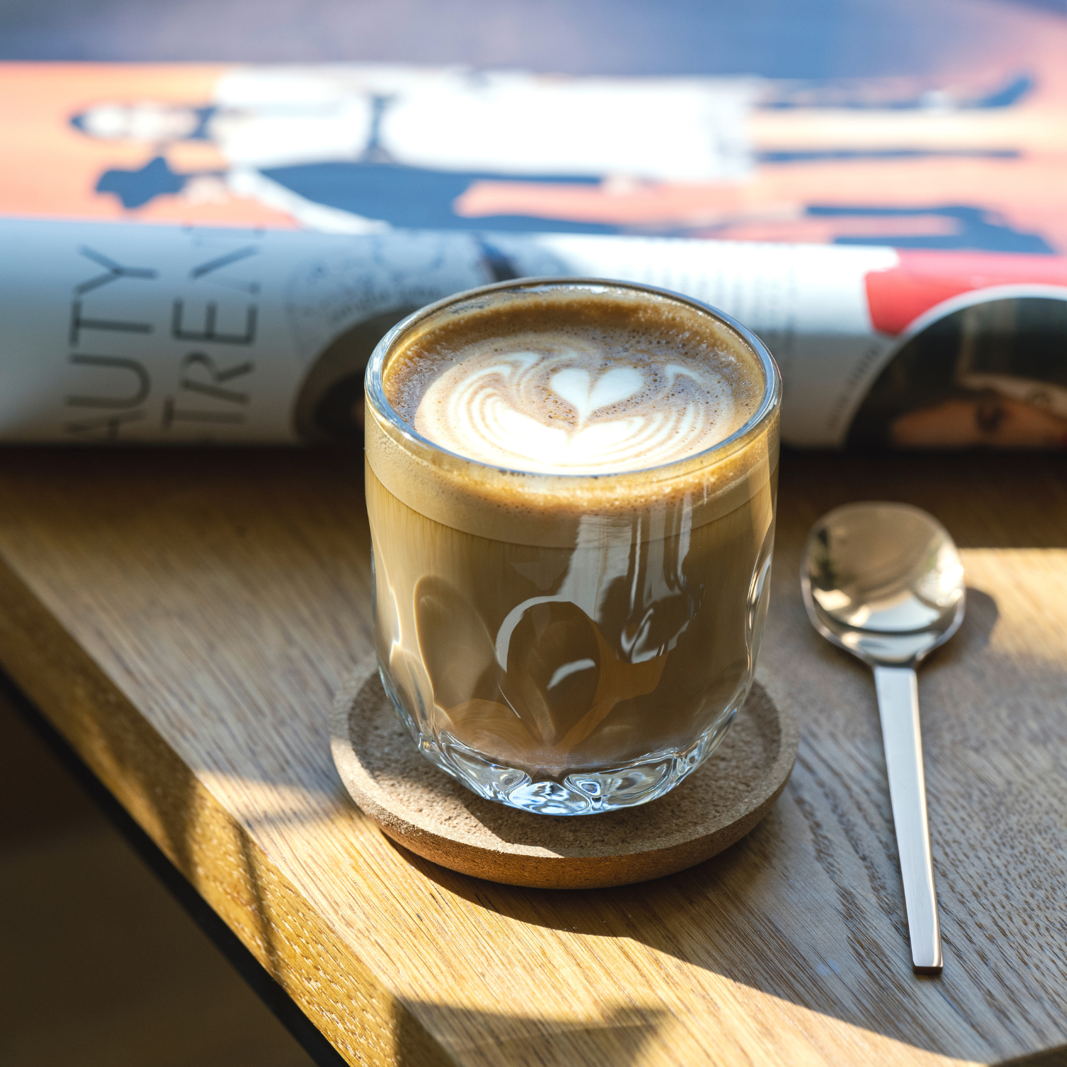 Ein Glas mit Latte Art in Herzform steht auf einem Korkuntersetzer auf einem Holztisch, daneben liegt ein Löffel vor einem aufgeschlagenen Magazin.