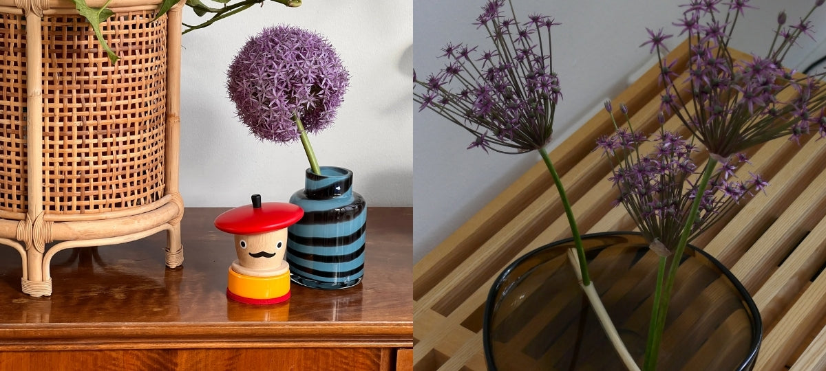 Dekorative Geschenkideen: Rattan-Lampenschirm, bunte Holzfigur, gestreifte Vase mit lila Allium-Blüte und Glas mit violetten Wildblumen