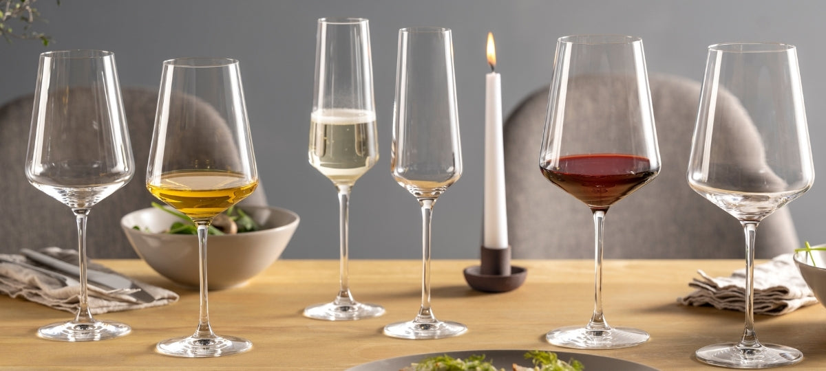 Elegante Weingläser in verschiedenen Formen gefüllt mit Wein und Wasser auf Holztisch, mit Kerze und grauem Hintergrund