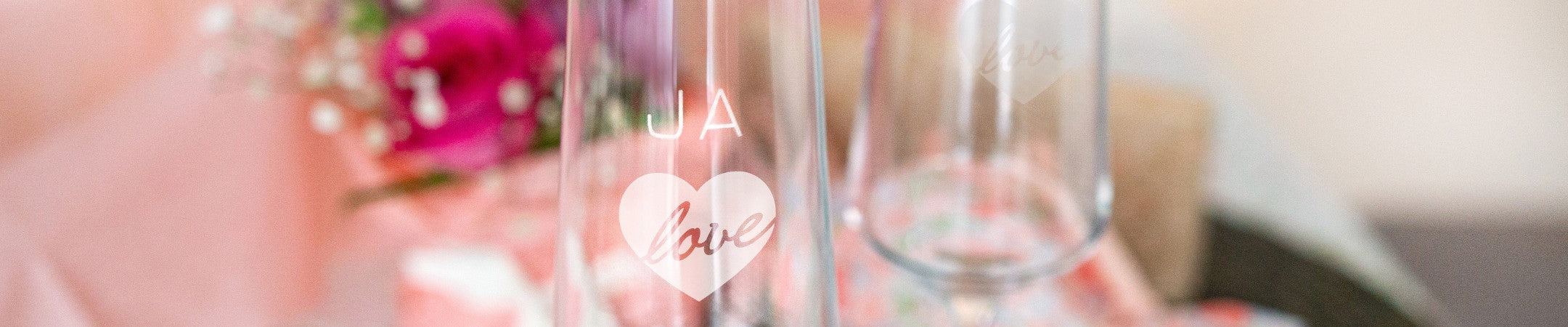 Zwei LEONARDO Sektgläser mit Gravur „JA" Text und "love“ im Herzdesign, festlich inszeniert mit Blumen und Geschenken im Hintergrund.