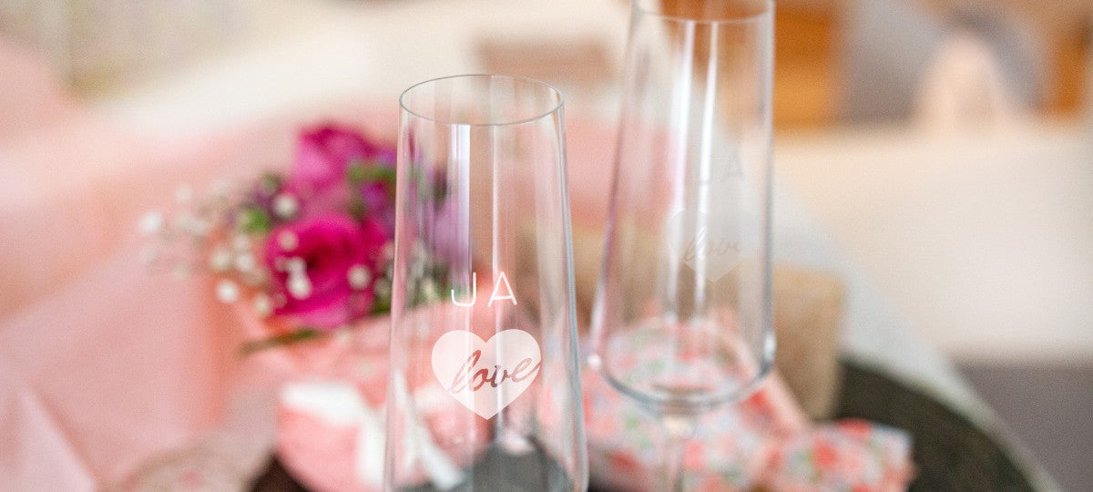 Zwei LEONARDO Sektgläser mit Gravur „JA" Text und "love“ im Herzdesign, festlich inszeniert mit Blumen und Geschenken im Hintergrund.