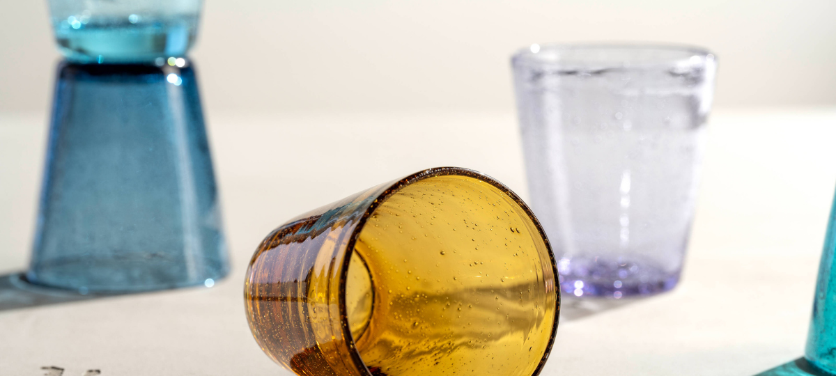 Ein bernsteinfarbenes Glas mit charakteristischen Luftblasen liegt quer im Mittelpunkt, während im Hintergrund weitere bunte Gläser in Türkis, Blau und Lila auf einem hellen Tisch arrangiert sind.