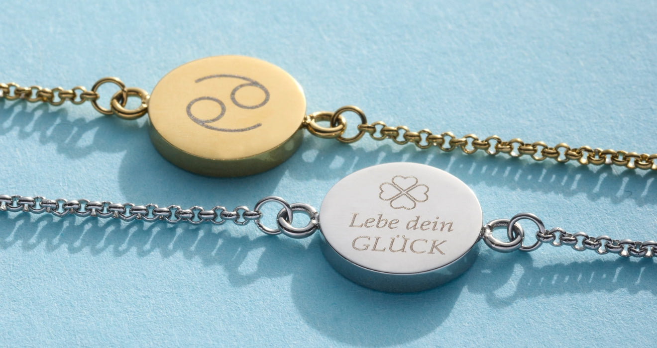 Zwei gravierte Armbänder auf hellblauem Hintergrund: eines in Gold mit dem Sternzeichen Krebs, das andere in Silber mit der Gravur „Lebe dein GLÜCK“ und einem Kleeblatt-Symbol.