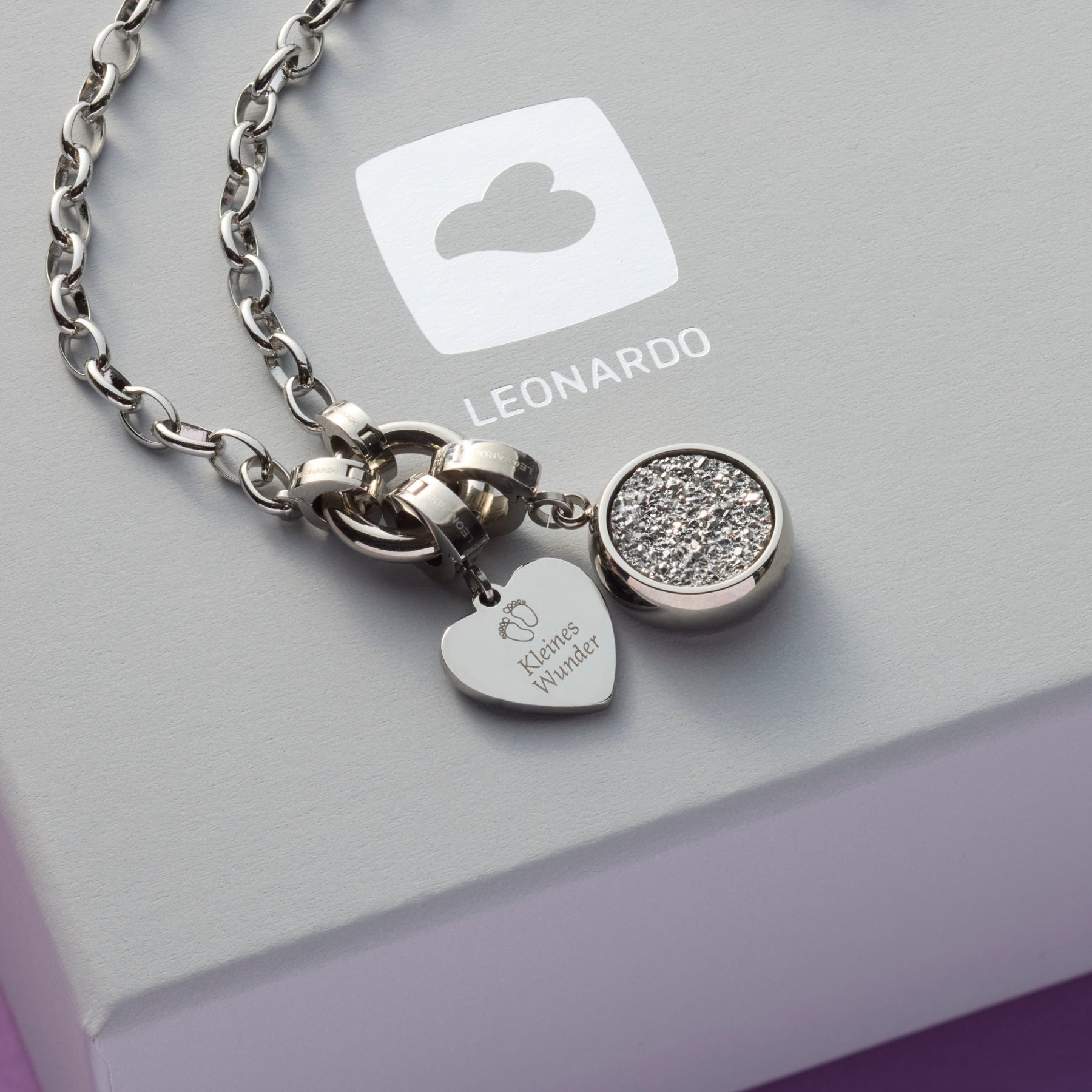 Zwei silberne Anhänger – ein rundes Element mit funkelnder Oberfläche und ein herzförmiges Plättchen mit der Gravur „Kleines Wunder“ – liegen auf einer grauen Geschenkbox mit LEONARDO-Logo.