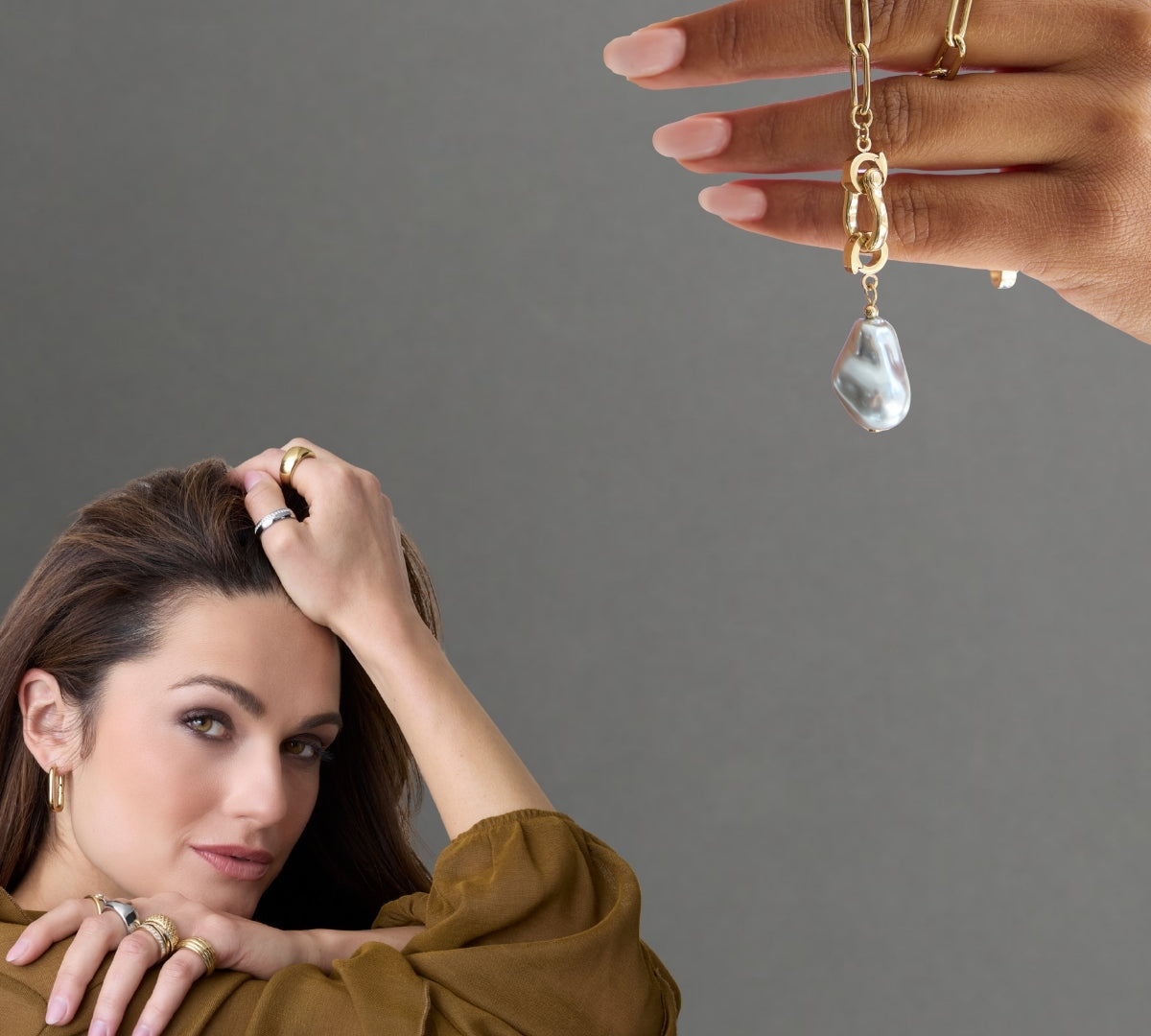 Eine Frau mit goldenen Creolen und mehreren Ringen in Silber und Gold legt entspannt ihre Hand ans Gesicht, daneben wird eine goldene Clip-Kette mit großer Barockperle in einer Hand präsentiert.